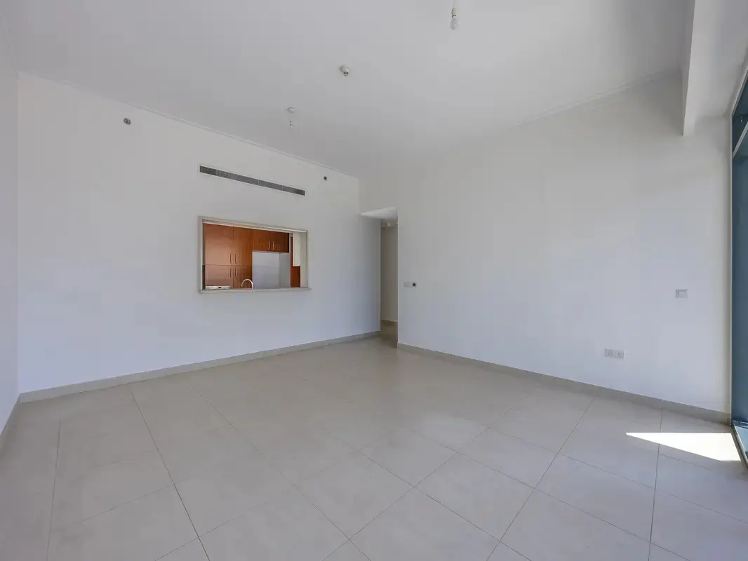 Empty room with white walls and beige tile floor. A kitchen pass-through reveals wood cabinets and a white refrigerator. Natural light enters from the right.