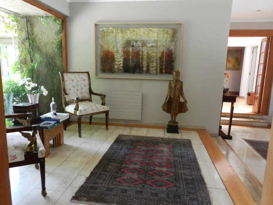 A foyer with a chair, a statue, and an abstract painting. A rug with red patterns lies on the tiled floor. A doorway leads to another room.