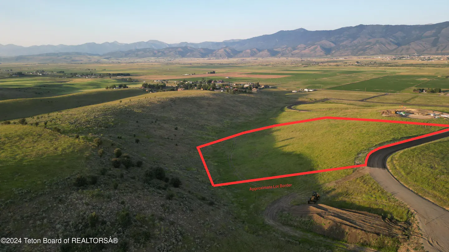 Aerial view of a grassy lot outlined in red, with mountains in the background. A road curves along the edge of the lot.