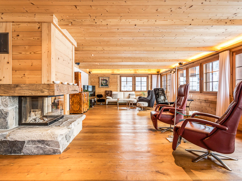 Spacious rustic living room with wood-paneled walls, a stone fireplace, and several leather armchairs on hardwood floors.