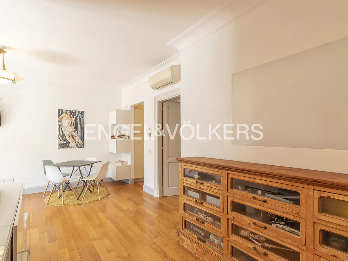 Bright living room with wood floors, white walls, and a large wooden cabinet. A dining table with chairs sits on a yellow rug. Artwork adorns the walls.
