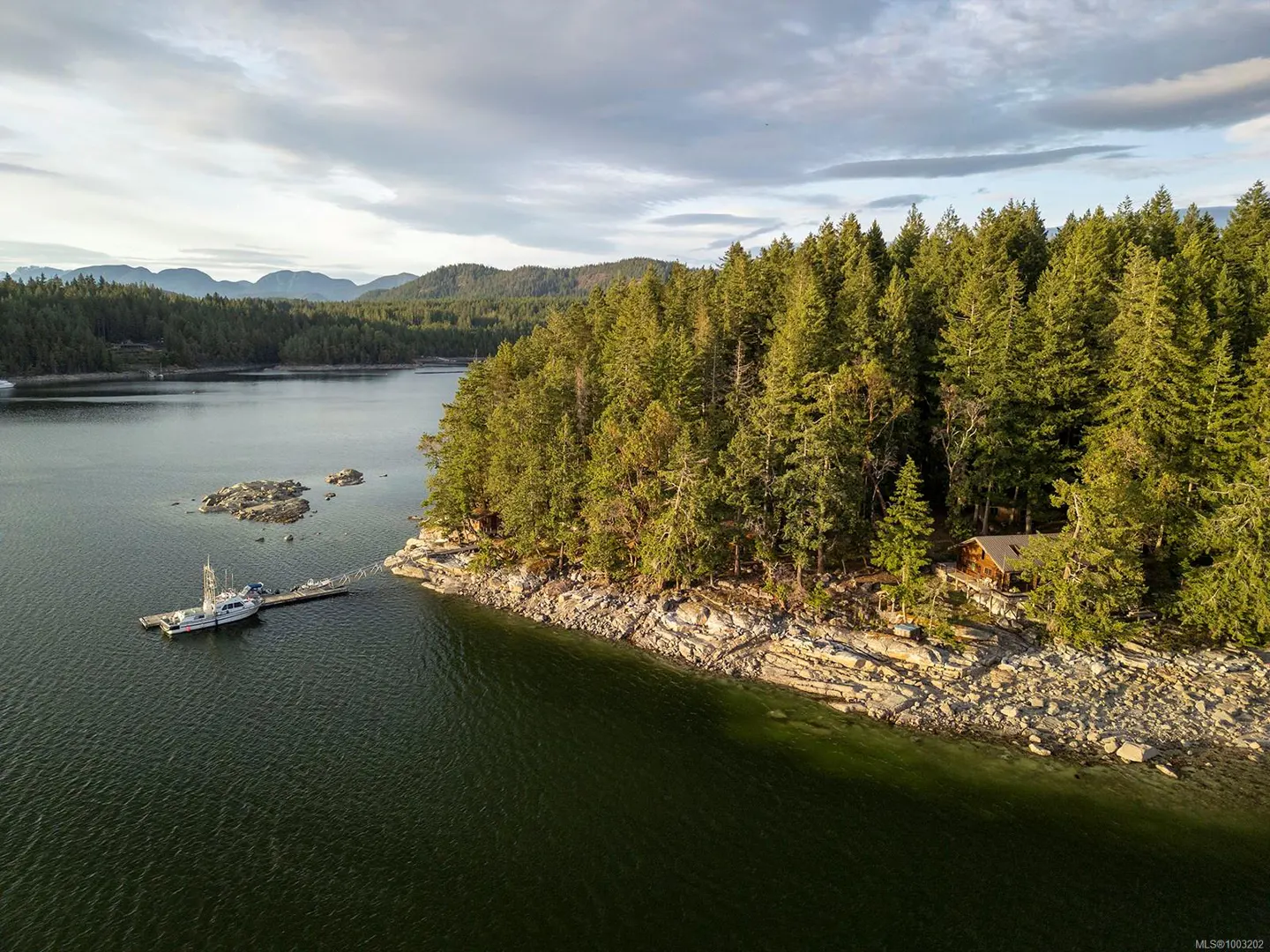 Aerial view of a boat docked at a rocky, tree-lined shore on a lake, with mountains in the distance.
