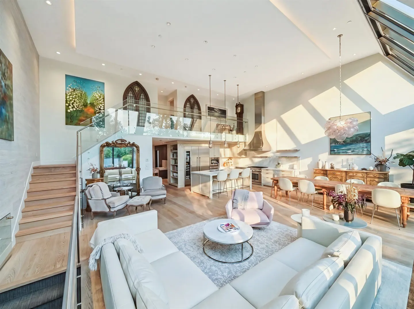 Bright, open-concept living space with white walls, wood floors, and a large white sectional sofa. A kitchen island and dining area are visible in the background. Natural light floods the room.