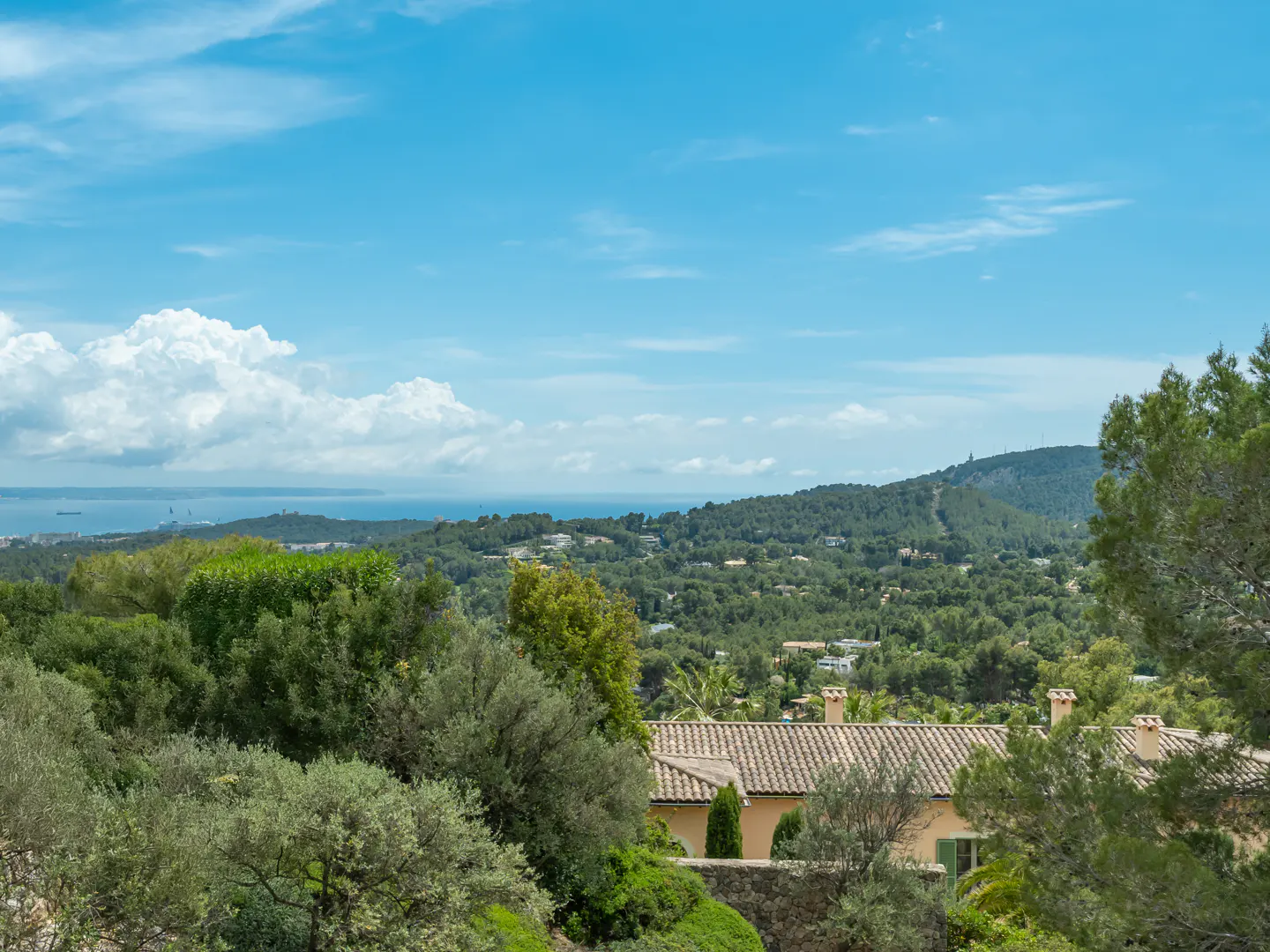 Scenic view of a tan house with a tiled roof, surrounded by green trees, with a blue sky and ocean in the background.