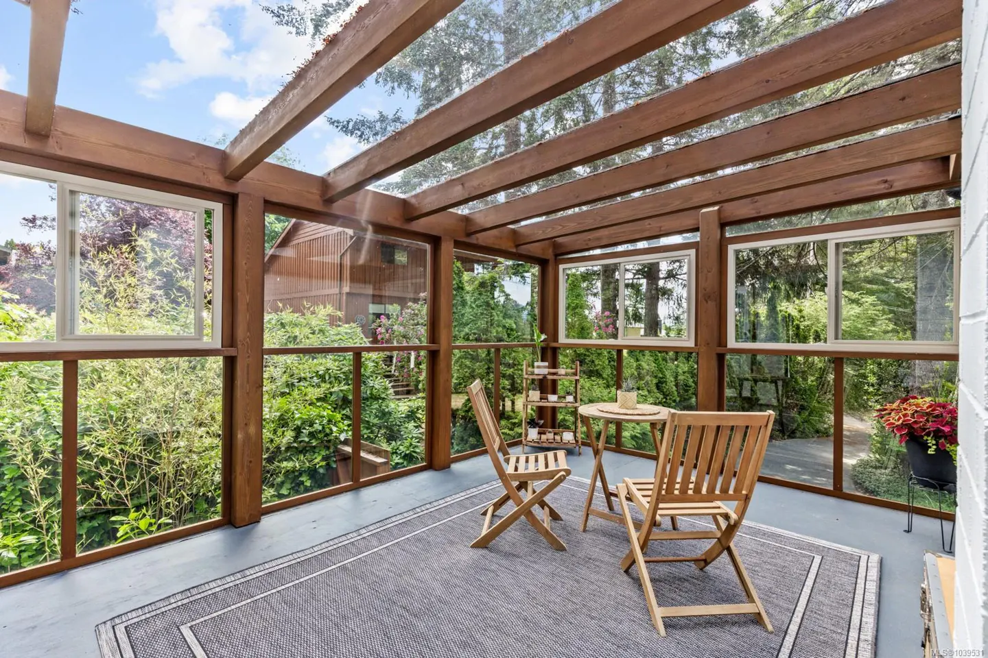 Sunroom with wooden beams, glass walls, a grey rug, and two wooden chairs around a small table overlooking a garden.