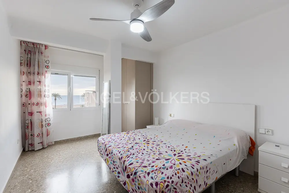 Bright bedroom with a double bed, patterned bedding, ceiling fan, and a window view of the sea and palm trees.