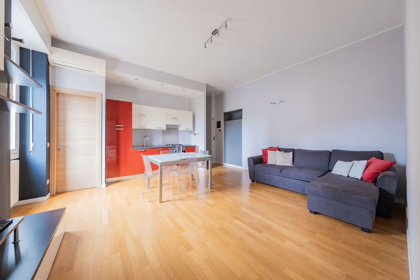 Open-concept apartment with wood floors, gray walls, and a gray sofa with red pillows. A white table and chairs sit near red and white kitchen cabinets.