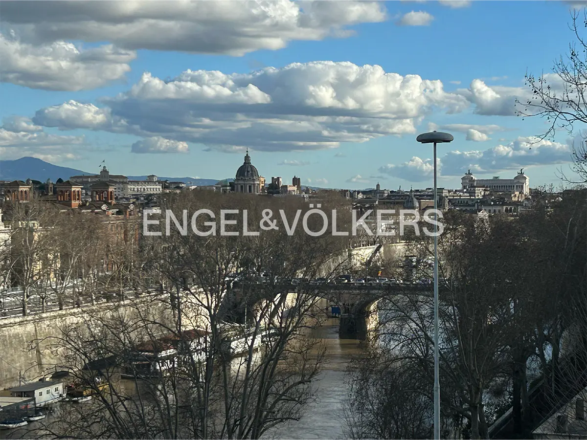 Panoramic view of the Tiber River in Rome with historic buildings, a stone bridge, and a cloudy blue sky.