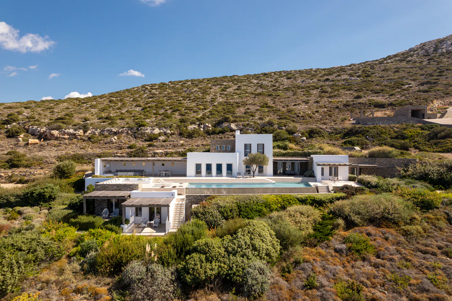 Modern white villa with a swimming pool nestled in a rugged, sun-drenched hillside landscape under a clear blue sky.
