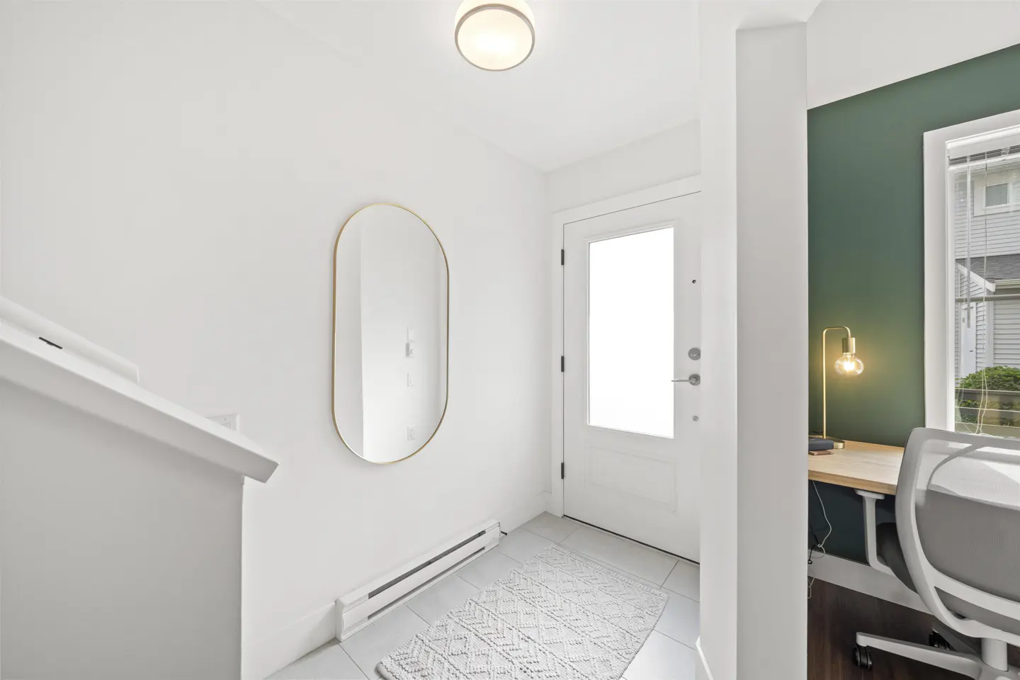 Bright entryway with white walls, tile floor, and a white door. A gold-framed mirror hangs on the wall, and a white rug lies on the floor. A desk area is visible through an opening.