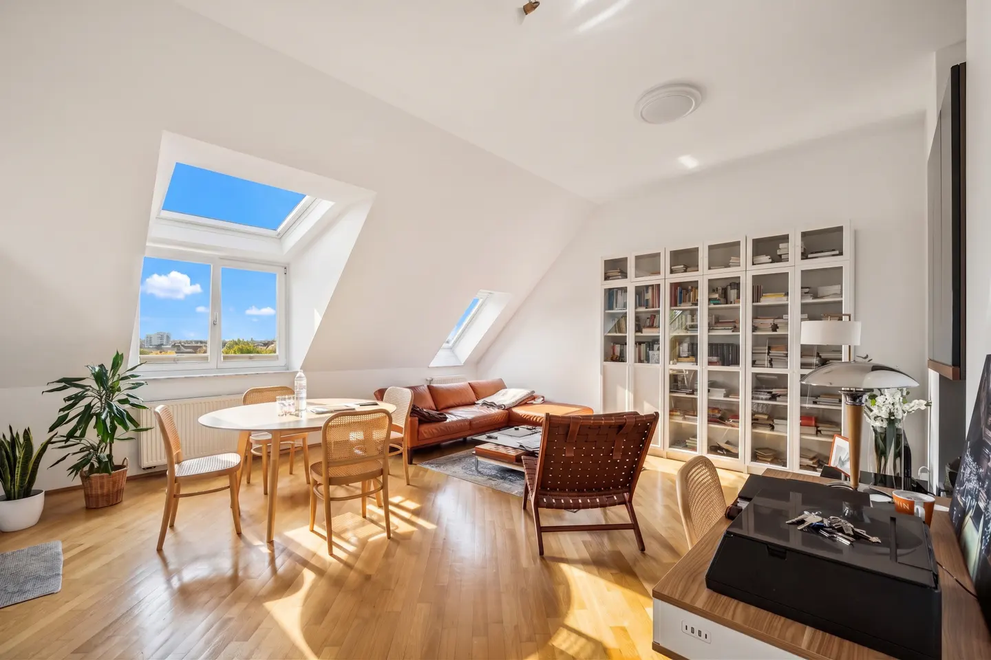 Bright attic apartment with wood floors, white walls, skylights, a brown leather sofa, and a white bookcase.