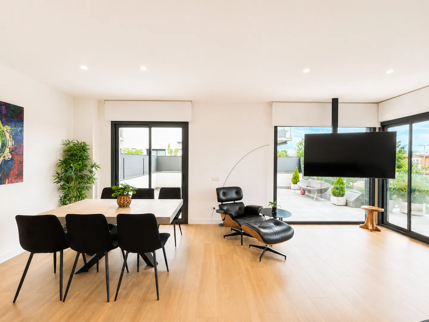 Modern dining area with black chairs, a lounge chair with ottoman, and a large TV overlooking a terrace with glass doors.