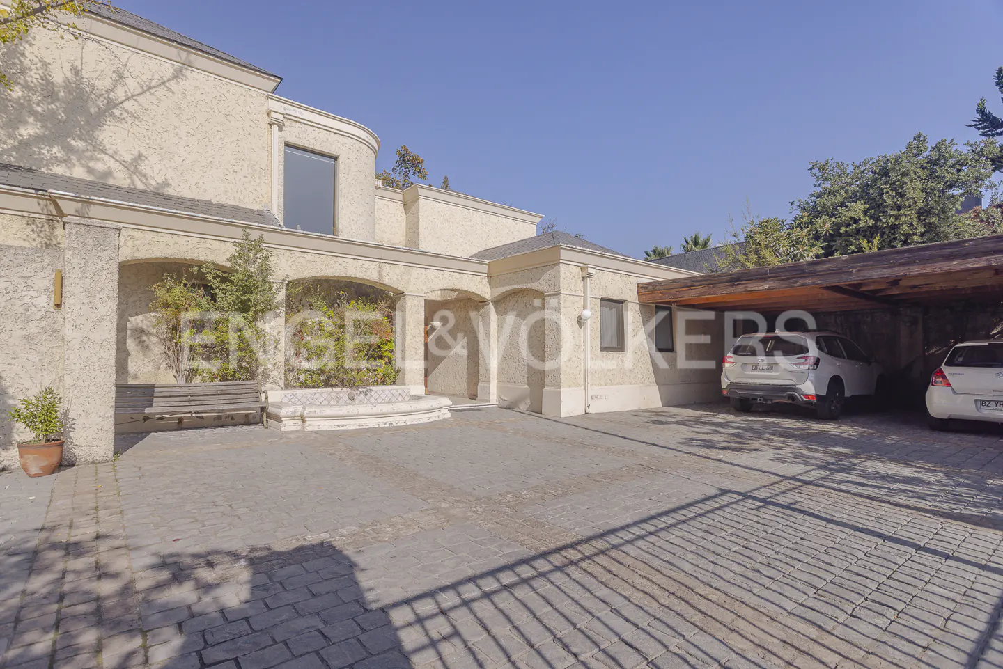 Exterior view of a beige stucco house with a stone driveway and two cars parked under a wooden carport.