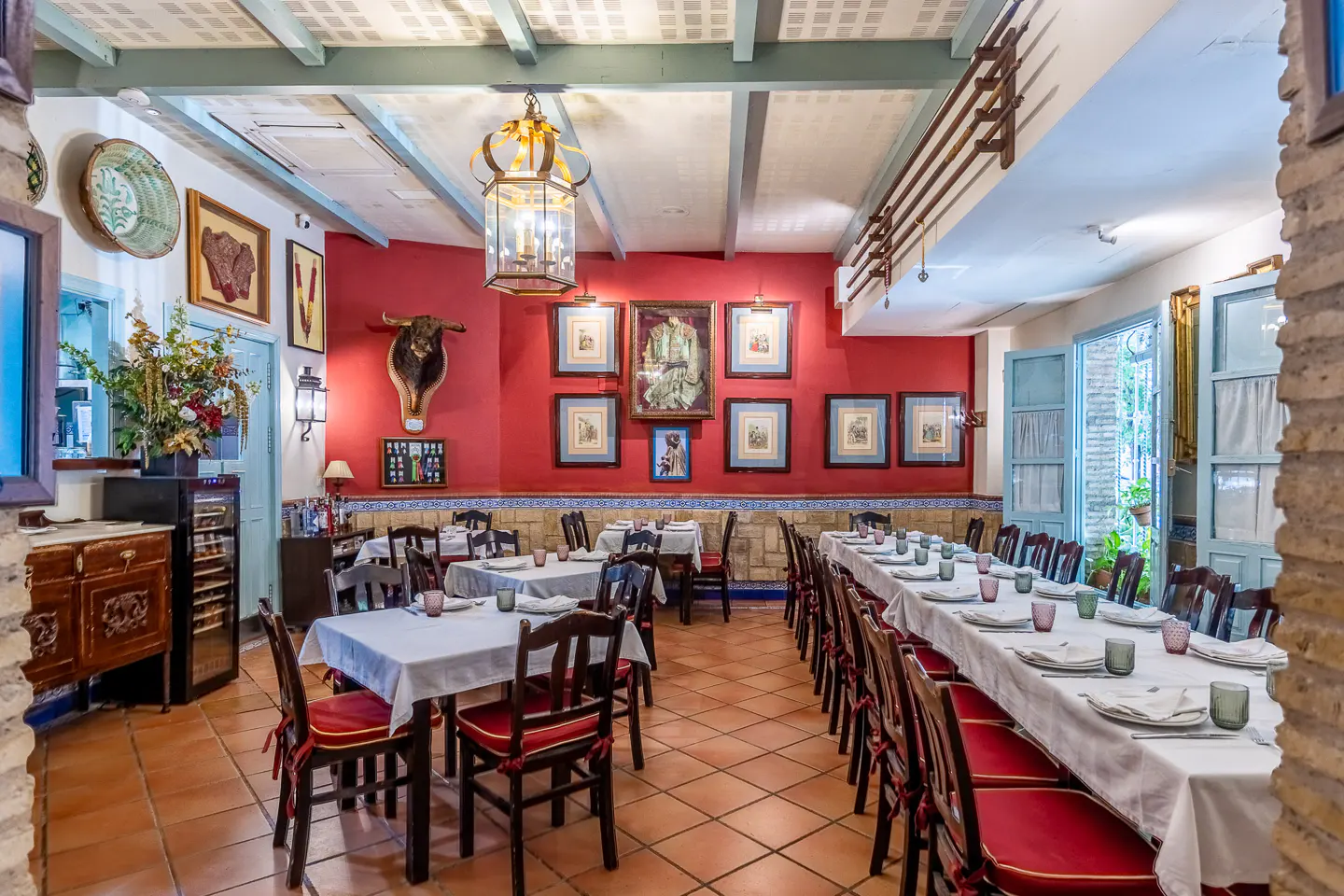 A rustic restaurant interior with red walls, terracotta floors, white-clothed tables, and chairs with red cushions.