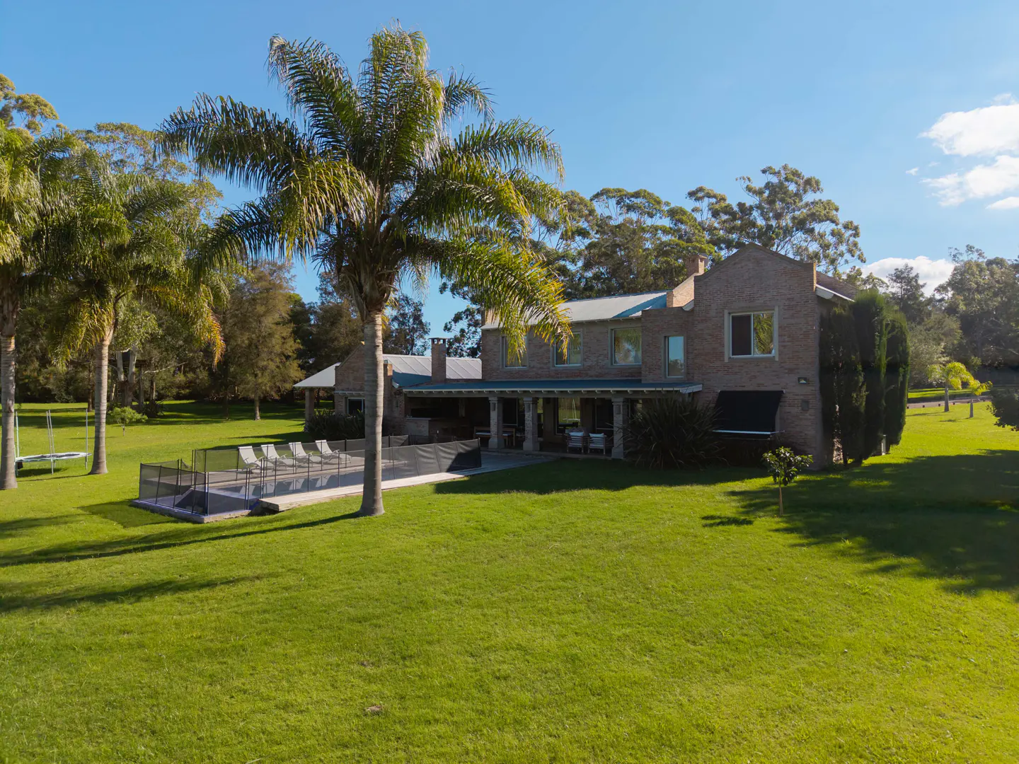 Exterior view of a two-story brick house with a pool, palm trees, and a large green lawn under a blue sky.