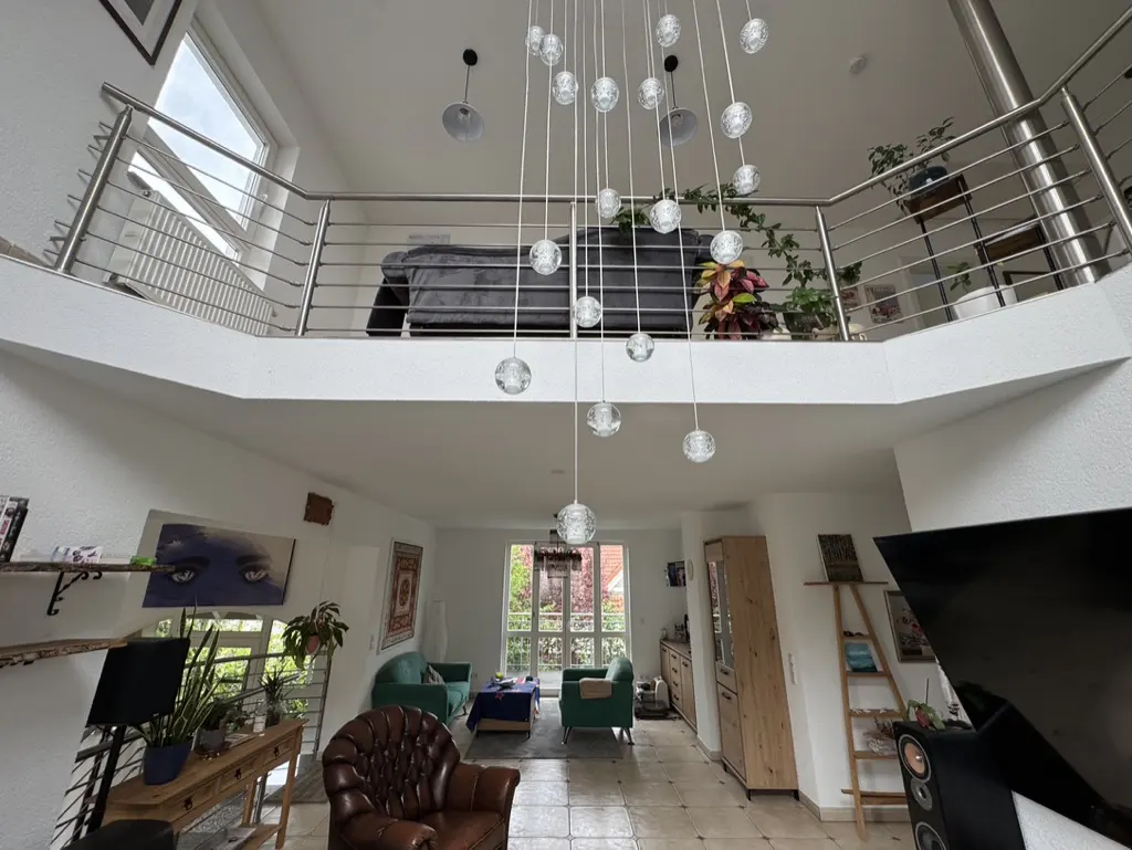 A bright, open-concept living room with a second-floor loft. A chandelier of glass orbs hangs from the ceiling. The room features a brown leather chair and green sofas.