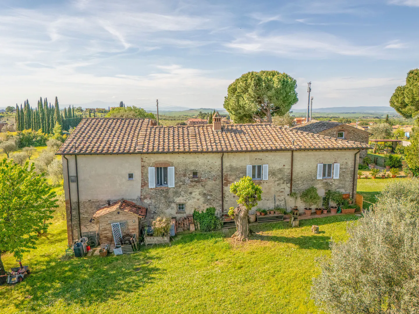 Aerial view of a rustic, tan-colored house with a red tile roof, white shutters, and green lawn in a rural setting.
