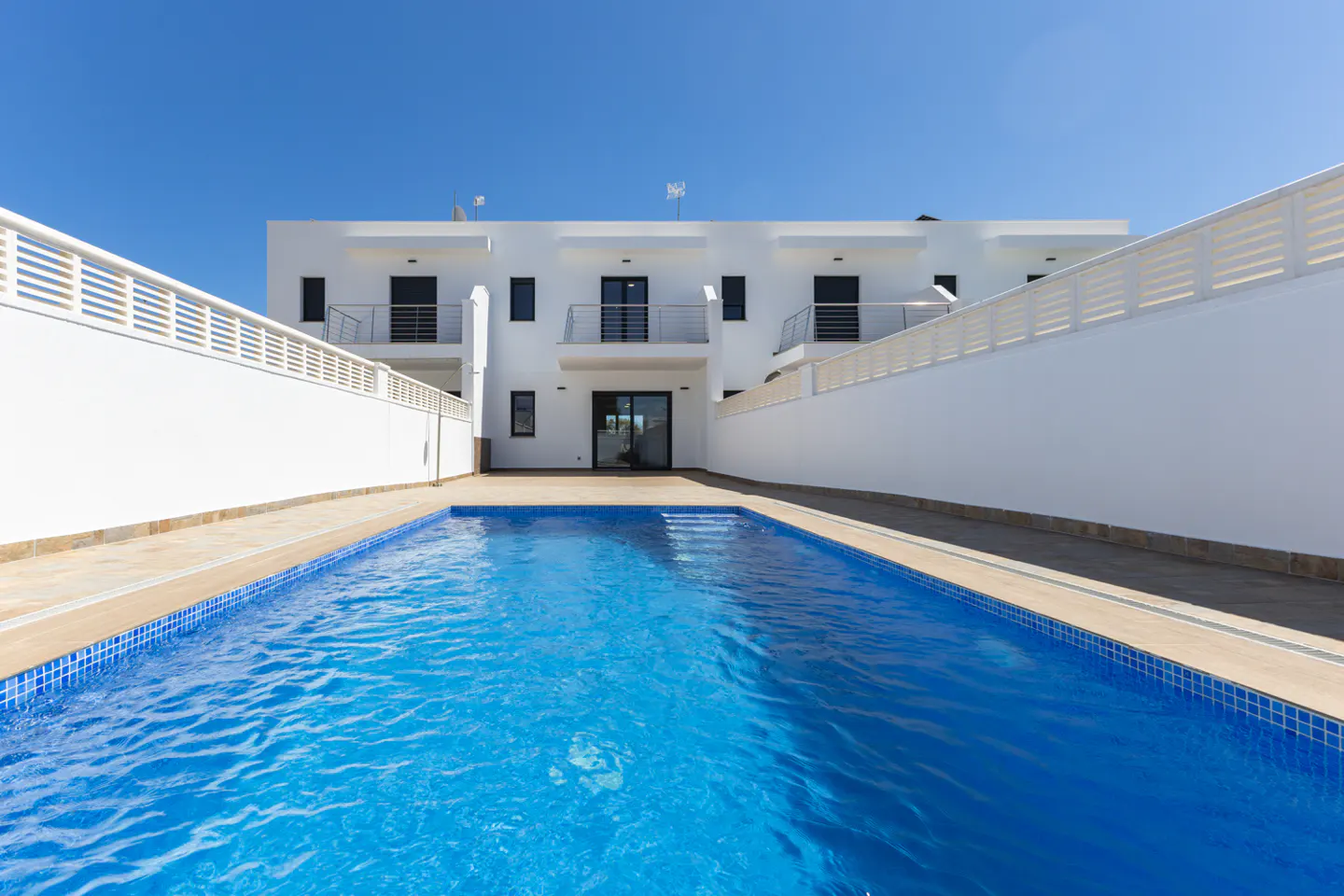 A modern white building with balconies overlooks a blue swimming pool under a clear blue sky.