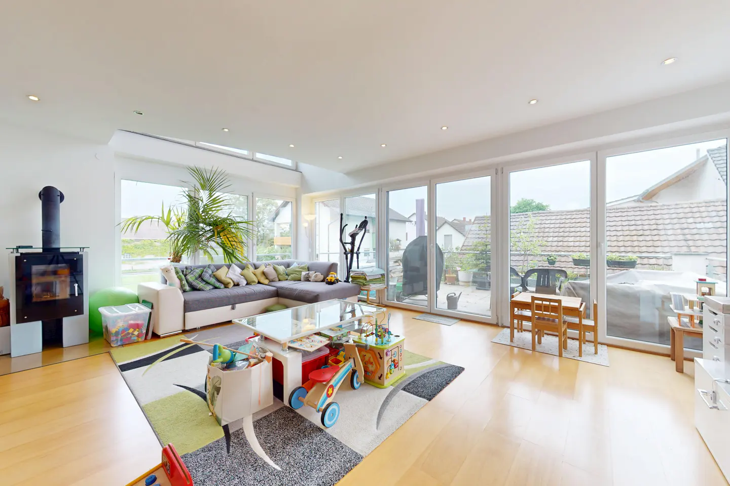 Bright living room with wood floors, a gray sectional sofa, and a modern fireplace. Toys are scattered on a colorful rug. Large windows lead to a patio.
