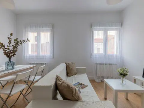 Bright living room with white walls, wood floors, and two windows with sheer curtains. A white sofa with pillows and a table with chairs are visible.