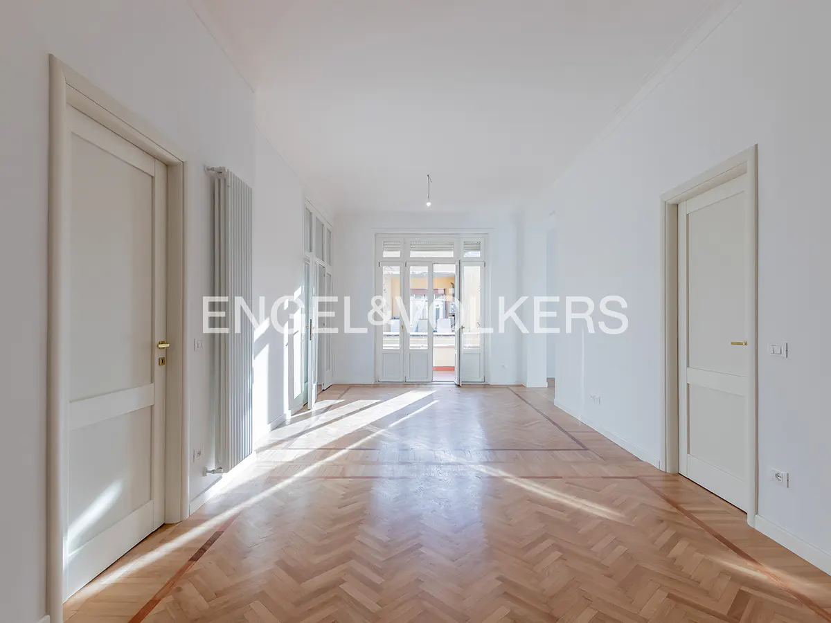 Bright, empty room with herringbone wood floors, white walls, and two white doors. A radiator is on the left wall. Double doors lead to a balcony.