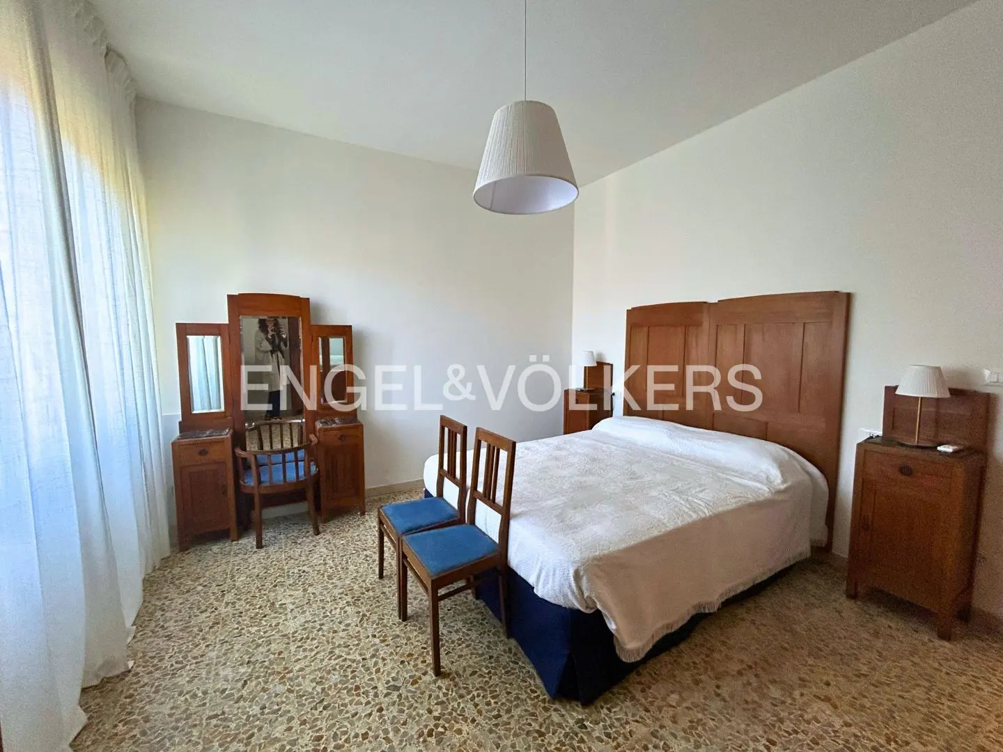 Bedroom with a wooden bed frame, two chairs, and a vanity with a mirror. A white lampshade hangs from the ceiling. The floor is tiled.