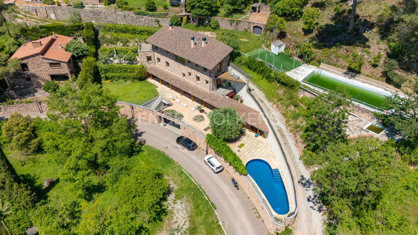 Aerial view of a stone house with a red tile roof, pool, and cars on a winding driveway. Lush green trees surround the property.