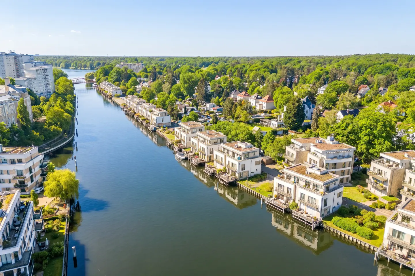Aerial view of modern white waterfront homes lining a canal, surrounded by lush green trees under a clear blue sky.
