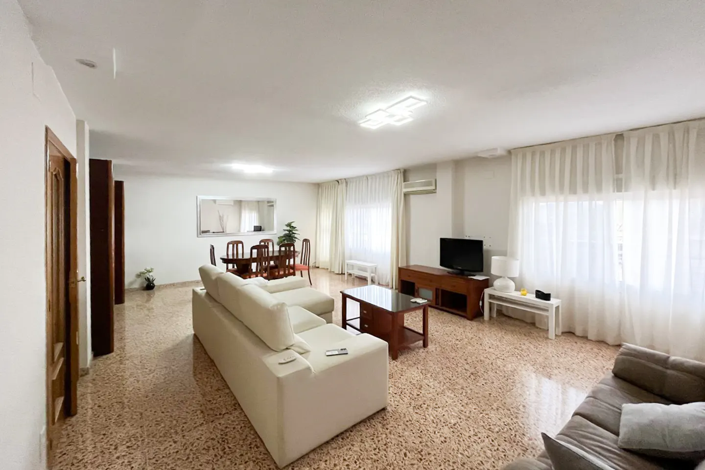 Bright living room with white walls, speckled floor, and white sectional sofa. Dining table, TV, and sheer white curtains are also visible.