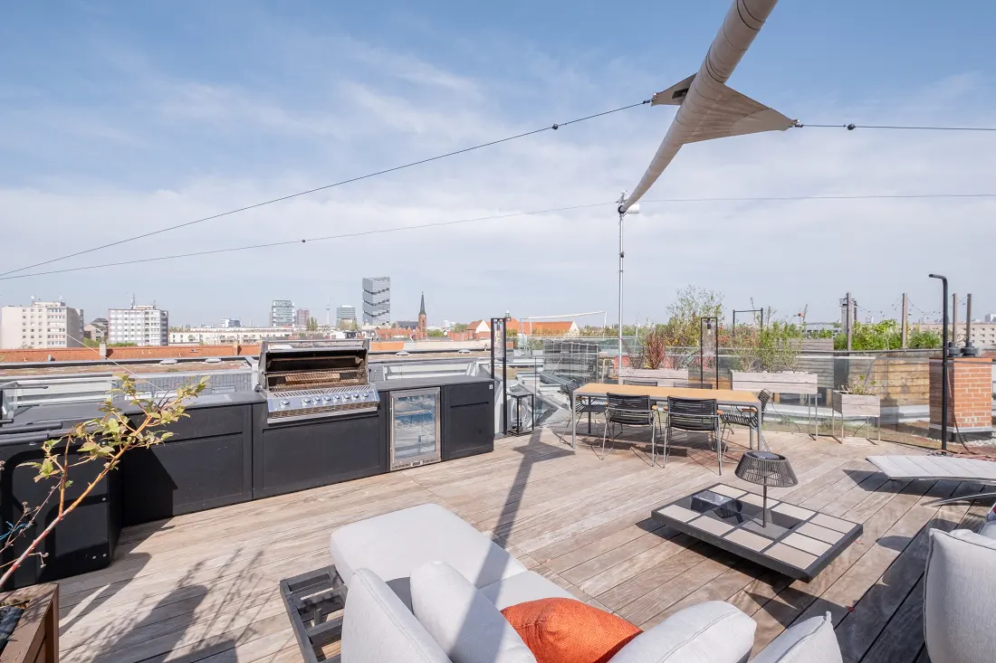 Rooftop patio with wood decking, grill, dining table, lounge chairs, and city views under a bright sky.