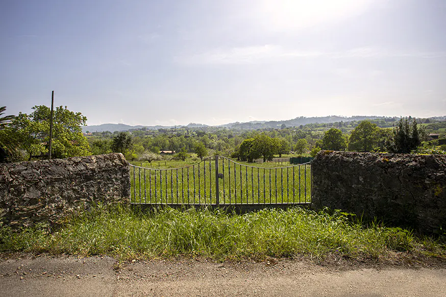 Iron gate between stone walls, leading to a green field with trees and distant hills under a blue sky.