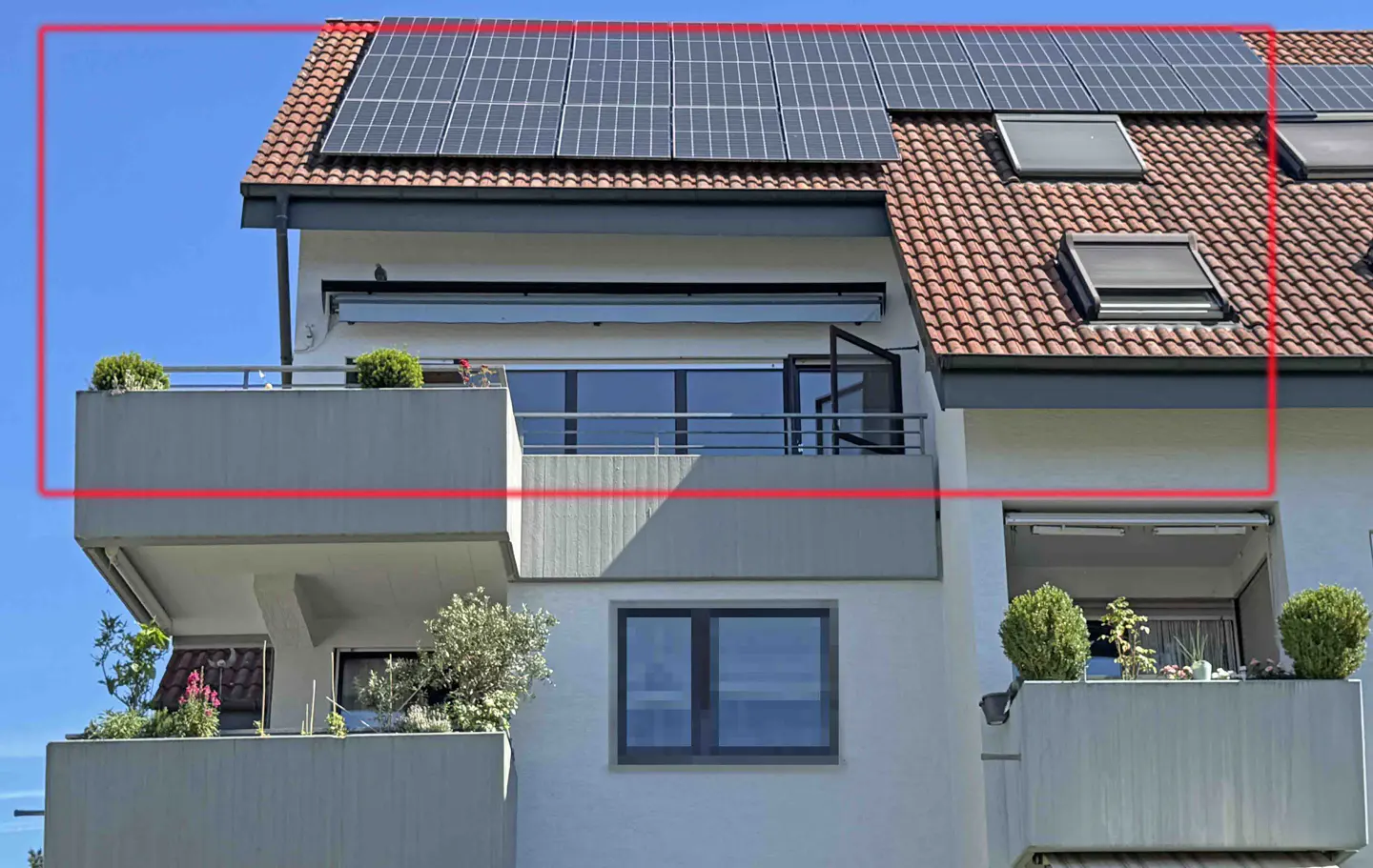 Modern apartment building exterior with solar panels on a tiled roof, a balcony with potted plants, and a clear blue sky.