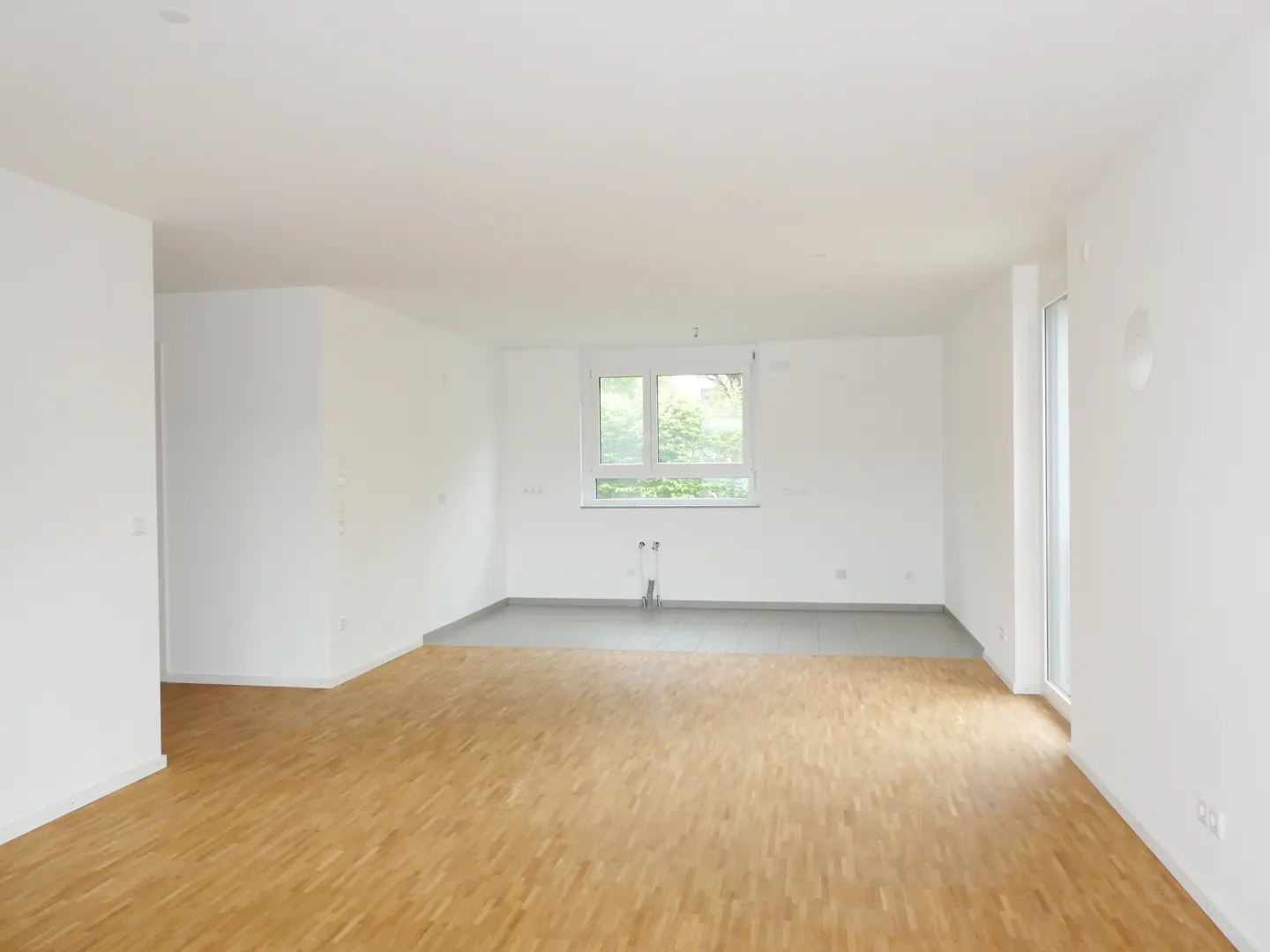 Bright, empty room with wood floors and white walls. A window lets in natural light, and a doorway leads to another room.