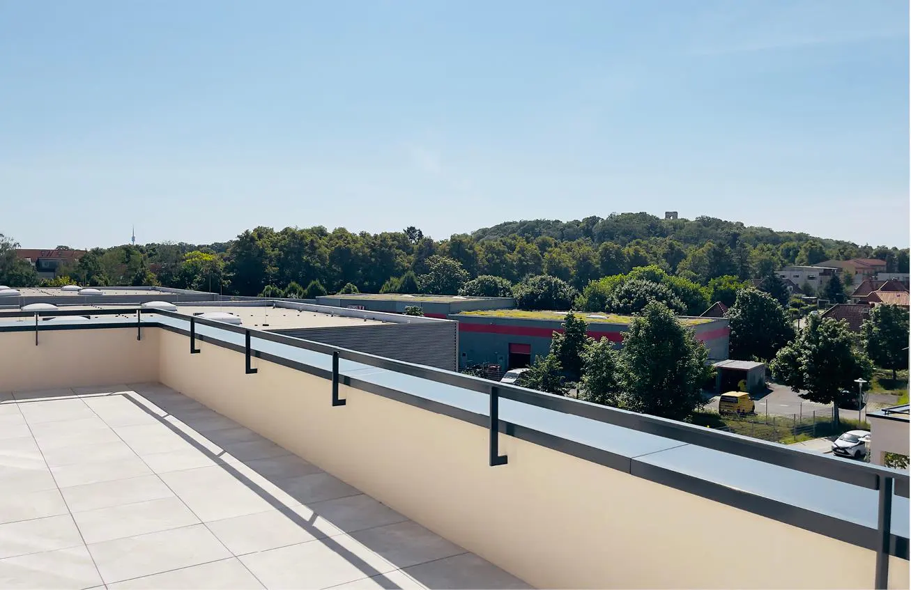 View from a rooftop terrace with a black railing, overlooking buildings, trees, and a distant hill under a clear blue sky.