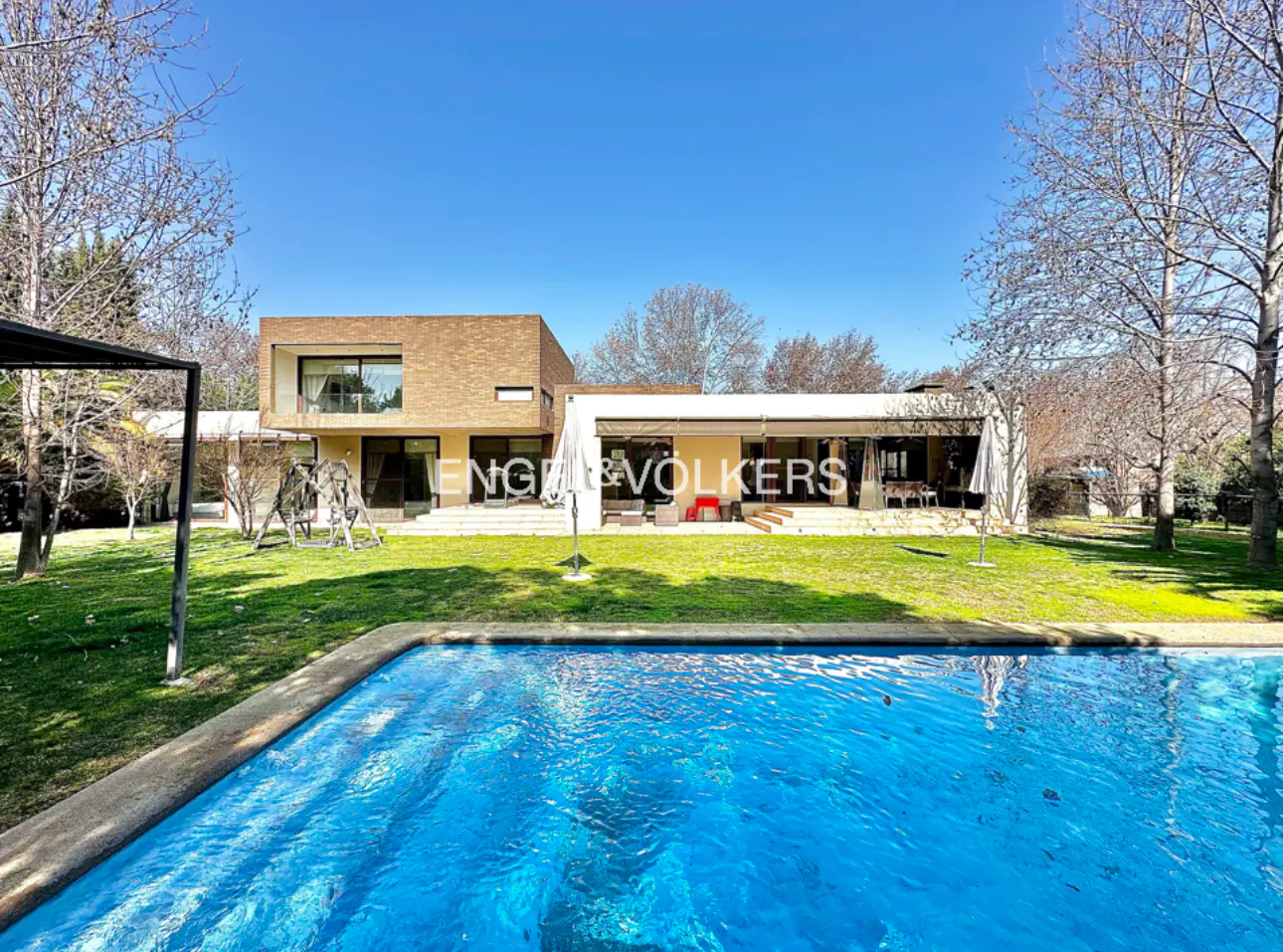 Exterior view of a modern two-story house with a blue swimming pool in the foreground. Green lawn and trees surround the property.