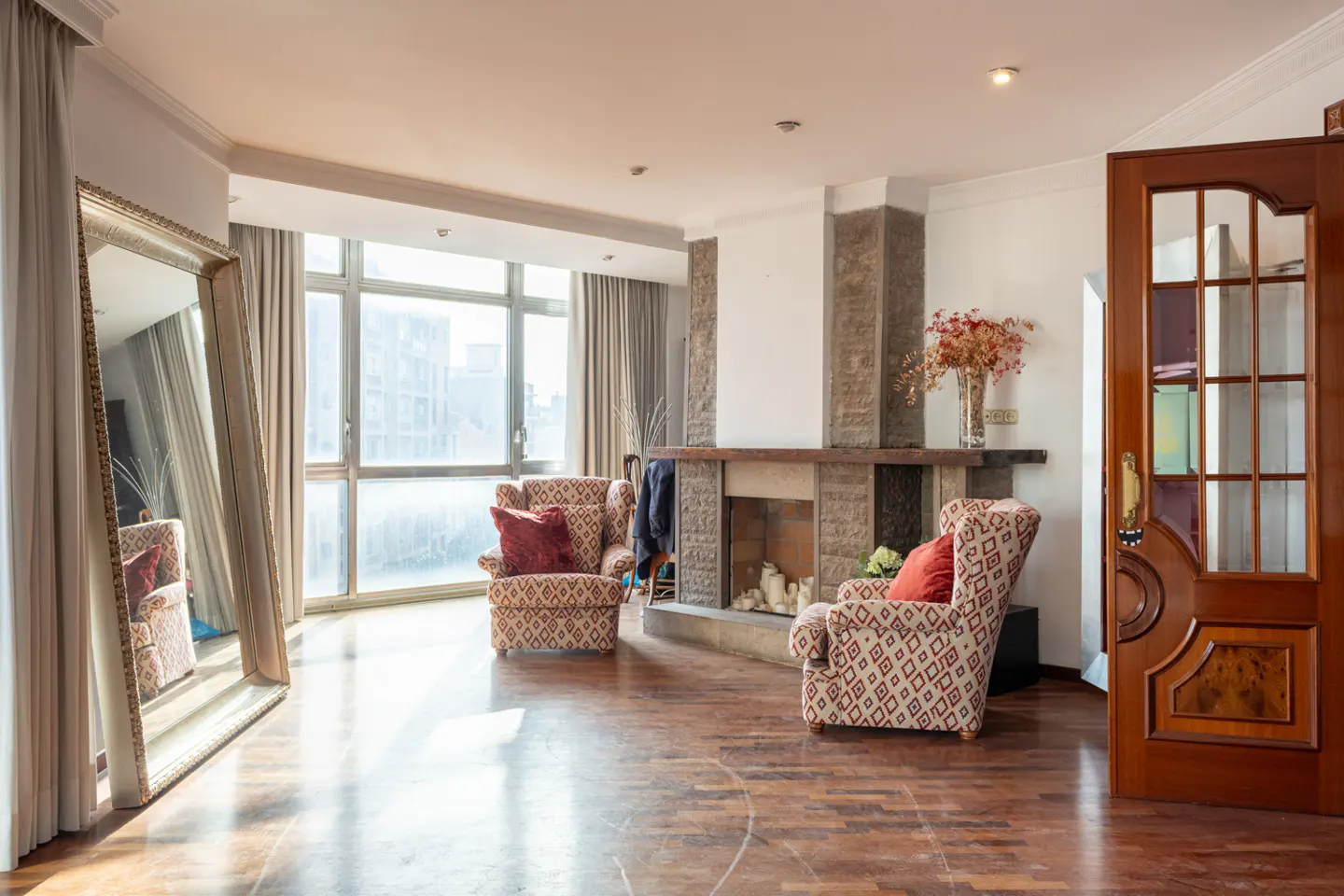 Living room with hardwood floors, stone fireplace, patterned armchairs, and a large gold-framed mirror by a window.