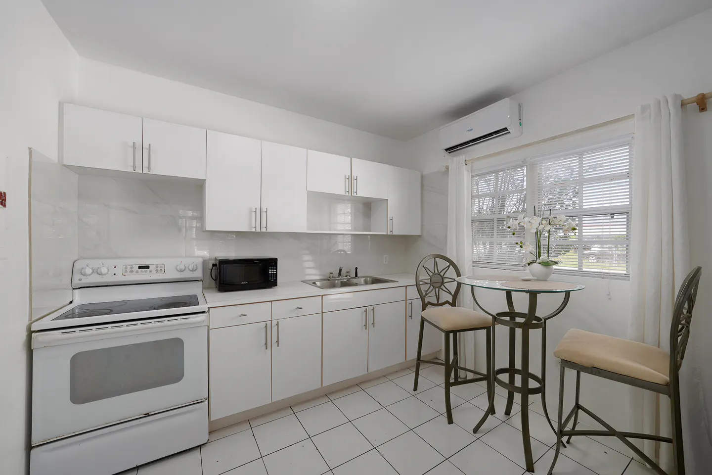 Bright, all-white kitchen with stove, microwave, cabinets, and a small table with two chairs near a window.