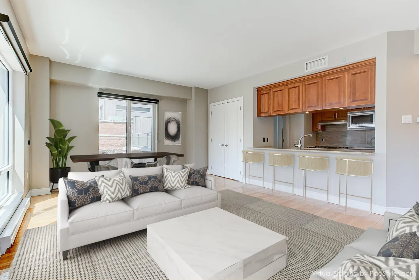 Bright, open-concept living space with a white sofa, coffee table, and bar stools at the kitchen counter. Large windows provide natural light.