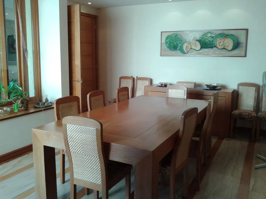 A dining room with a large wooden table surrounded by chairs with woven backs. A painting of green gourds hangs above a wooden sideboard.