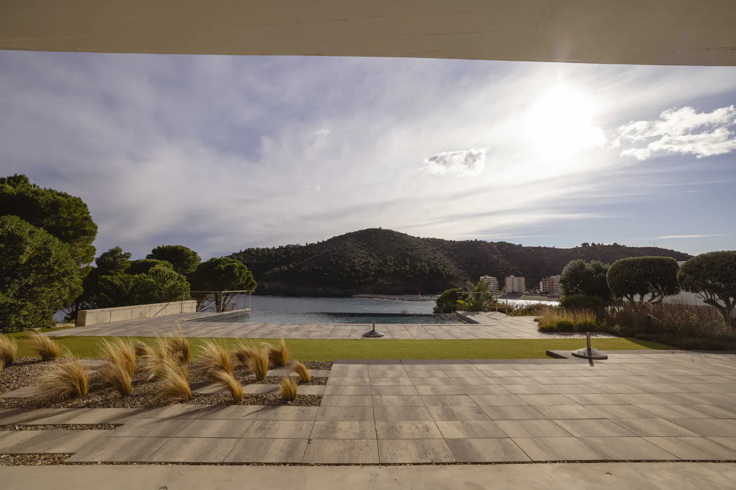 View from a patio with stone tiles, grass, and ornamental grass, overlooking a bay with a forested hill and buildings in the distance.