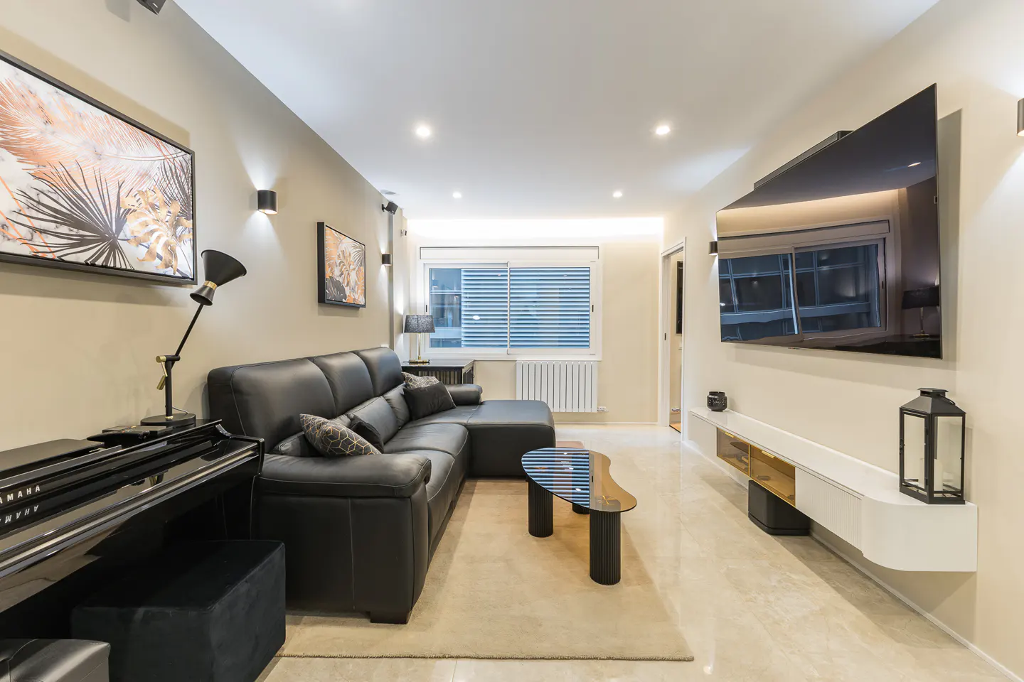 Living room with a black leather sofa, piano, and a large TV. The walls are beige, and there is a window with blinds.