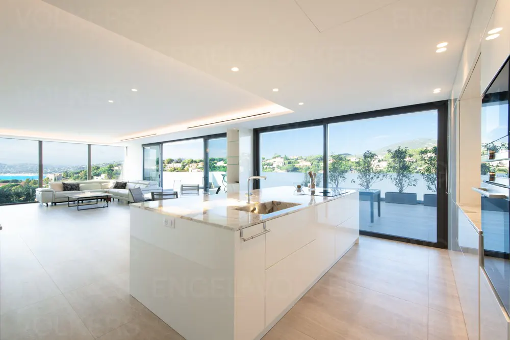 Bright, modern kitchen with a white island, marble countertop, and floor-to-ceiling windows overlooking a coastal view.