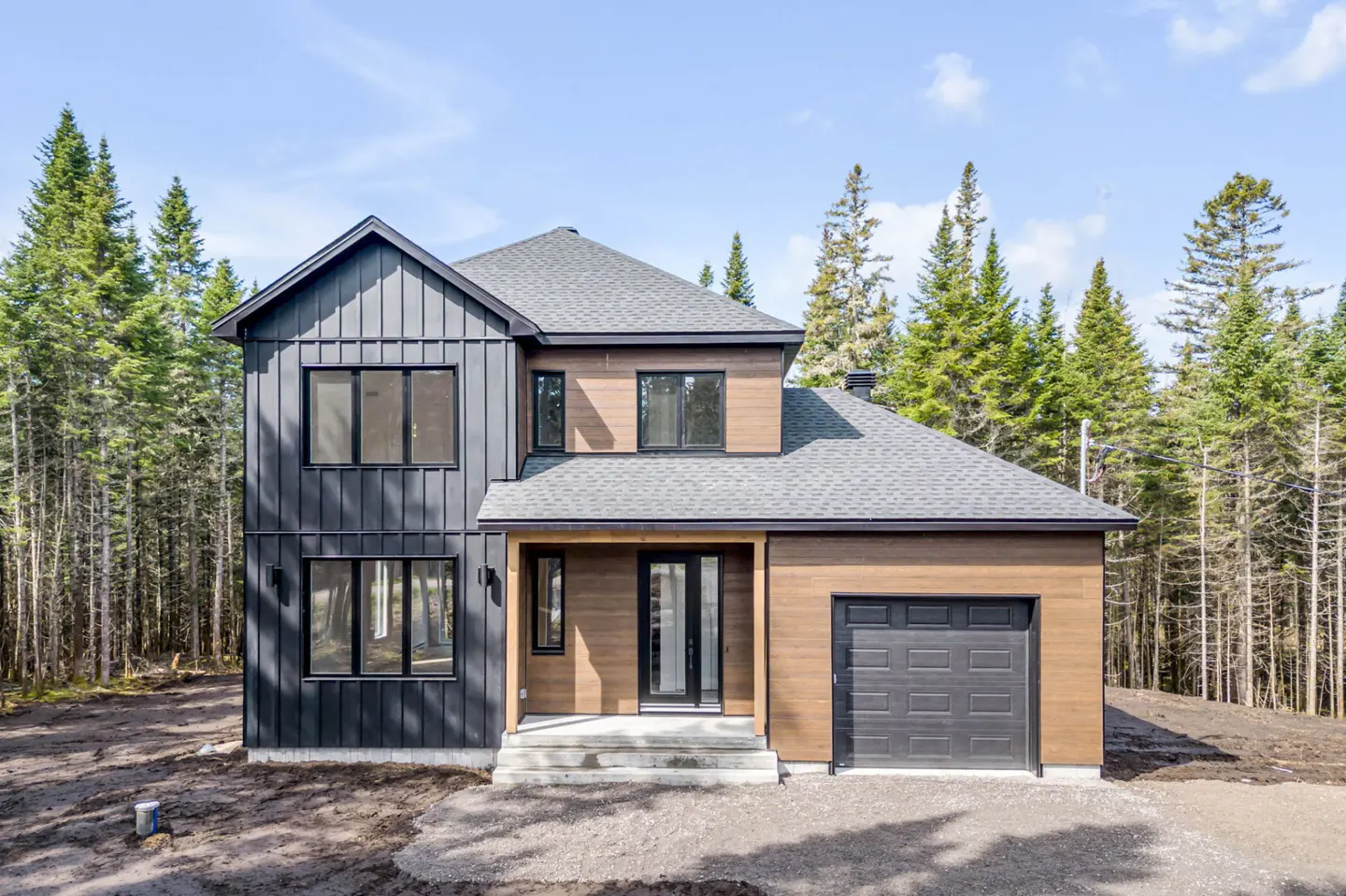 Two-story house with black and brown siding, gray roof, black trim, and a gray garage door. Tall green trees in the background.