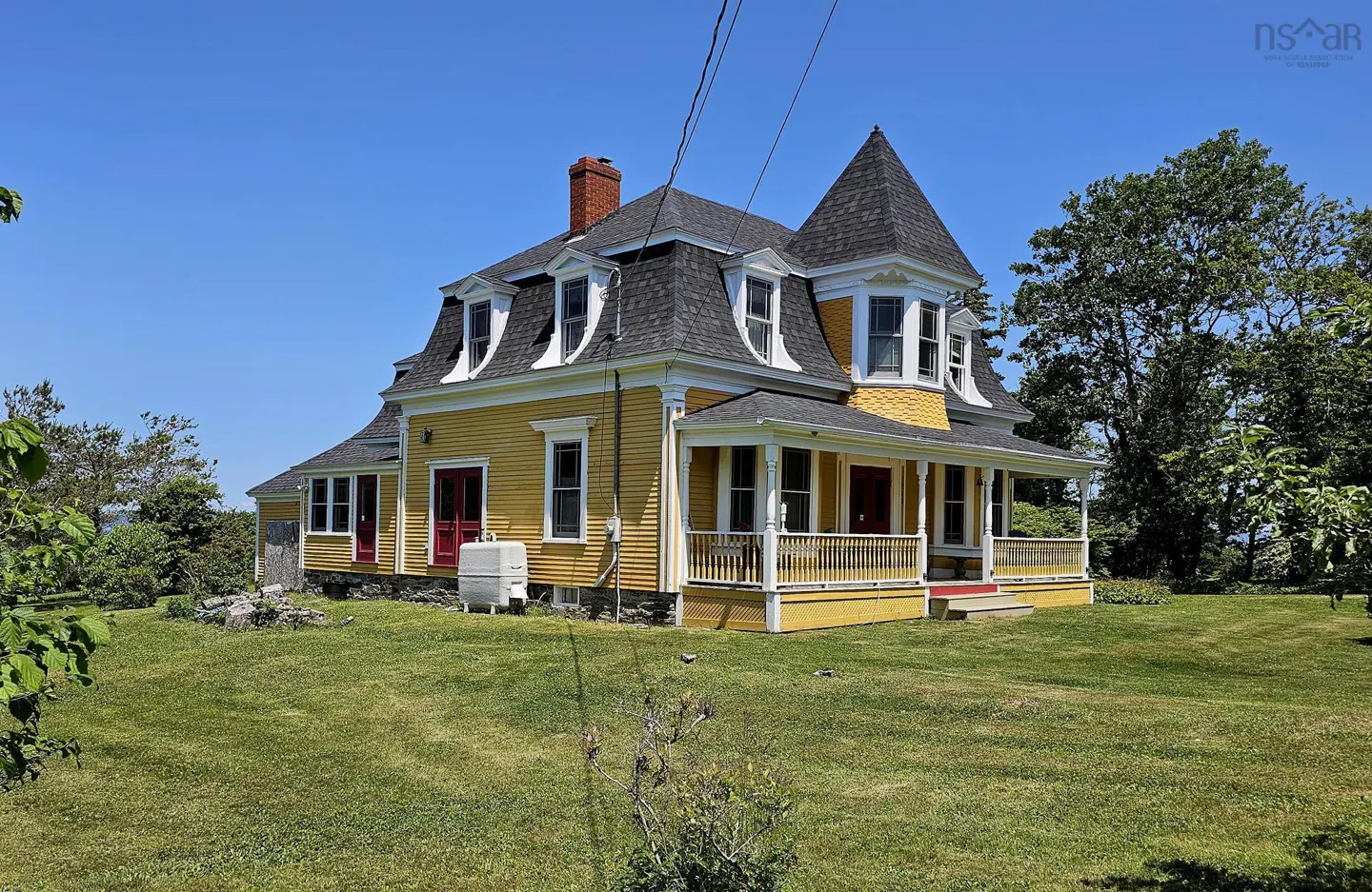 A yellow Victorian-style house with a turret, dark roof, and red doors, set on a grassy lawn under a clear blue sky.
