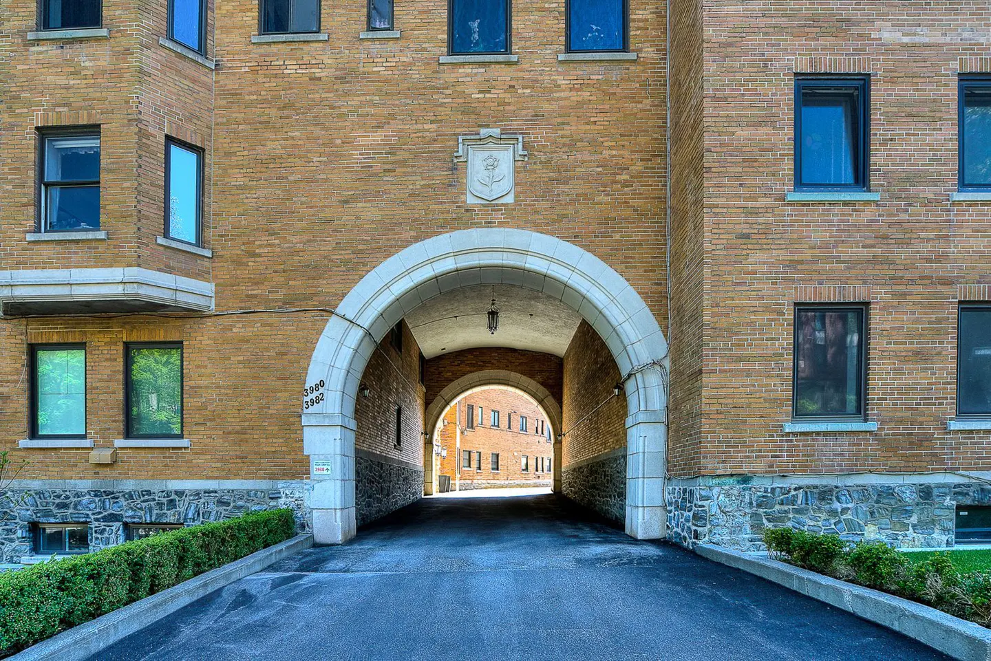 Exterior view of a brick apartment building with an arched entryway and a dark asphalt driveway.