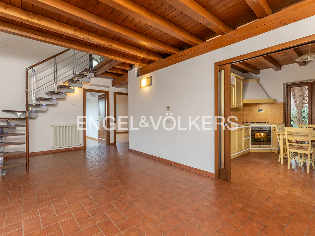 Open-concept living space with terracotta tile, wood-beamed ceiling, and a modern staircase. Kitchen visible through doorway.