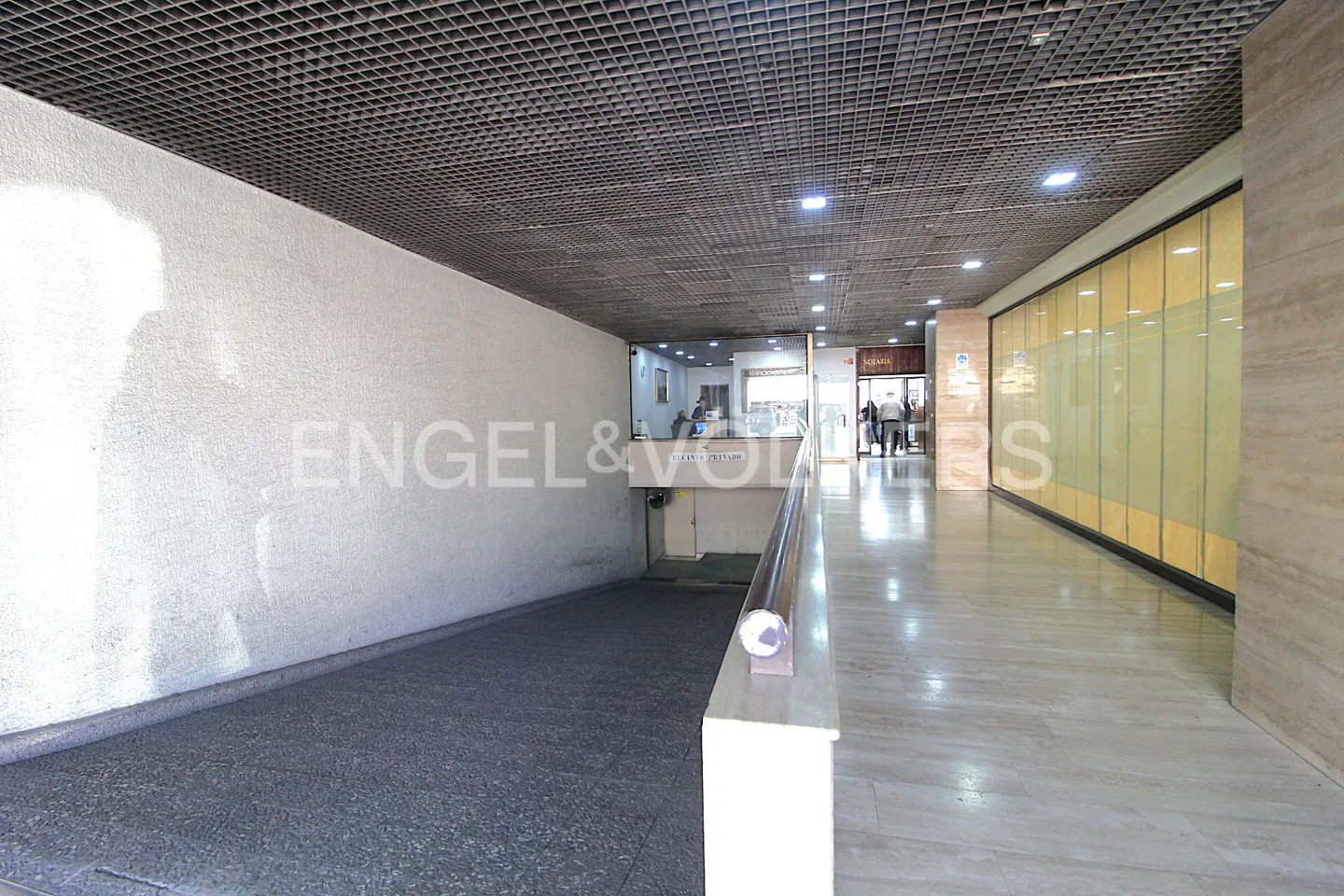 Interior view of a commercial building hallway with a ramp, marble floors, and a metal grid ceiling. A storefront is visible in the background.