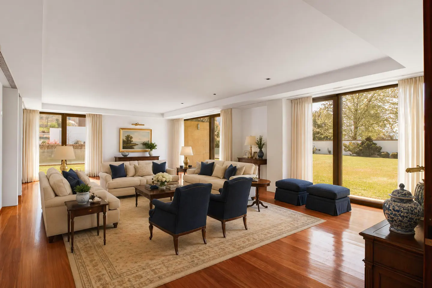 Bright living room with hardwood floors, beige sofas with blue pillows, and large windows overlooking a green lawn.