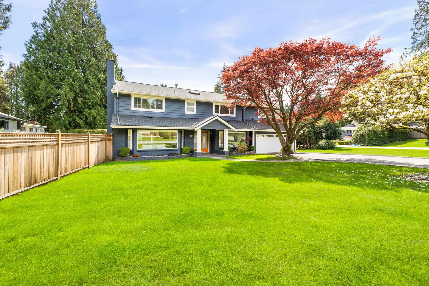 Two-story blue house with a green lawn, a red maple tree, and a wooden fence.