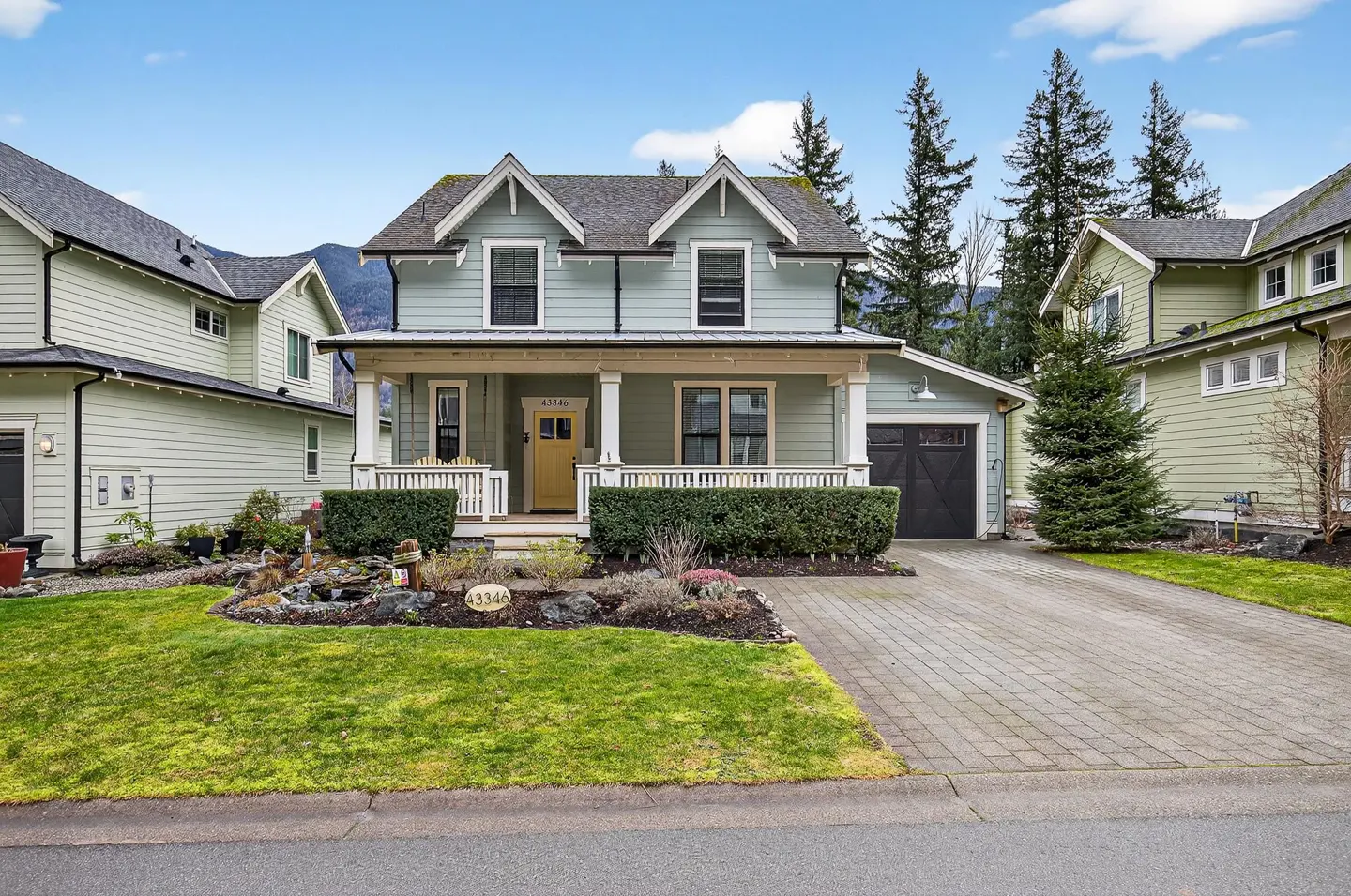 Two-story sage green house with a yellow front door, covered porch, paved driveway, and manicured front yard.